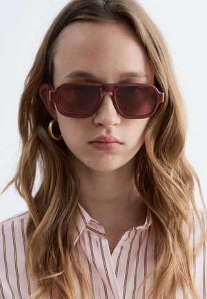 Jeune femme aux cheveux longs et ondulés portant de grandes lunettes de soleil teintées marron, des boucles d'oreilles créoles en or, et une chemise à rayures boutonnée.
