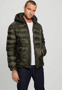 Olive green puffer jacket with a hood, quilted design, zip closure, and side pockets, worn over a white shirt and dark jeans.