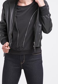 Svart bomberjacka i mjukt läder, med dragkedja fram och ribbade ärmar, lagd över en svart t-shirt med rund halsringning.