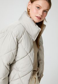 Chaqueta puffer beige ligera con cuello alto, patrón de rombos y cierre frontal con cremallera. Textura suave y lisa.