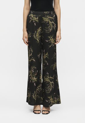 Γυναίκα φοράει μαύρο παντελόνι με φαρδιά πόδια και πράσινα και ροζ λουλουδάτα σχέδια, σε συνδυασμό με μαύρα σανδάλια με τακούνι και μαύρο τοπ.