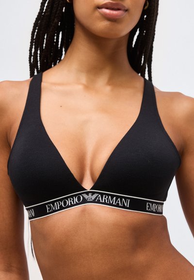 Γυναίκα που φοράει μαύρο bralette Emporio Armani με λωρίδα λογότυπου, δείχνοντας το άνω μέρος του κορμού και πλεξούδες στα μαλλιά της σε απλό φόντο.