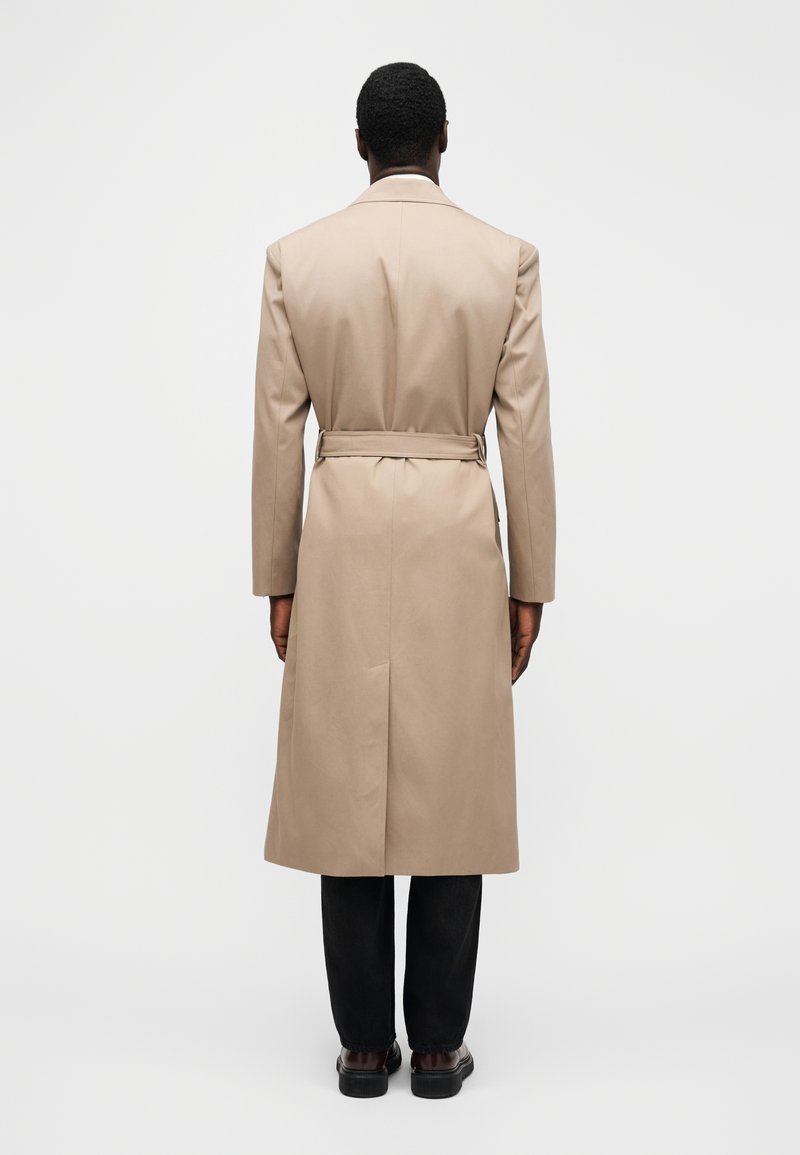 Trench-coat beige avec ceinture à la taille, manches longues et une fente à l'arrière. Le tissu semble lisse avec une silhouette nette et ajustée.