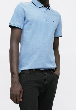 Mężczyzna w jasnoniebieskiej koszulce polo z krótkim rękawem z ciemnymi obszyciami i czarnych dżinsach, stojący z jedną ręką w kieszeni, na białym tle.