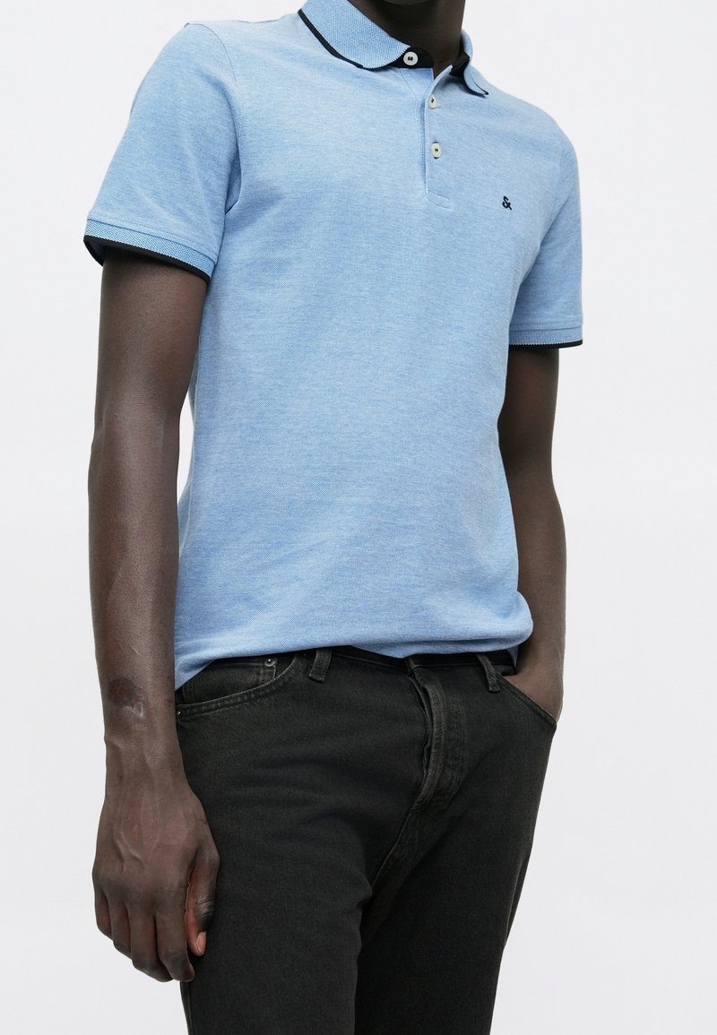 Homme portant un polo à manches courtes bleu clair avec des bordures foncées et un jean noir, debout avec une main dans la poche sur un fond blanc.