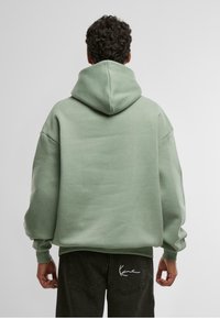 Prevelik zelen hoodie z veliko kapuco in rebrastimi manšetami. Gladka tekstura, sproščen dizajn. Črne hlače z belim vezenim detajlom na žepu.