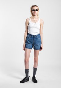 White ribbed tank top, blue denim shorts, gray socks, and black sneakers. Accessories include layered necklaces and small black sunglasses.