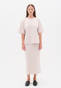 Kvinde med langt brunt hår, stående iført en løs lysebeige top, matchende lang nederdel og sorte slip-on sko på hvid baggrund.