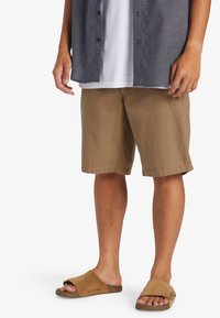 Pantalones cortos ligeros de color marrón con un ajuste relajado. Combinados con sandalias deslizantes de color beige. Capas con una camisa de botones gris sobre una camiseta blanca.