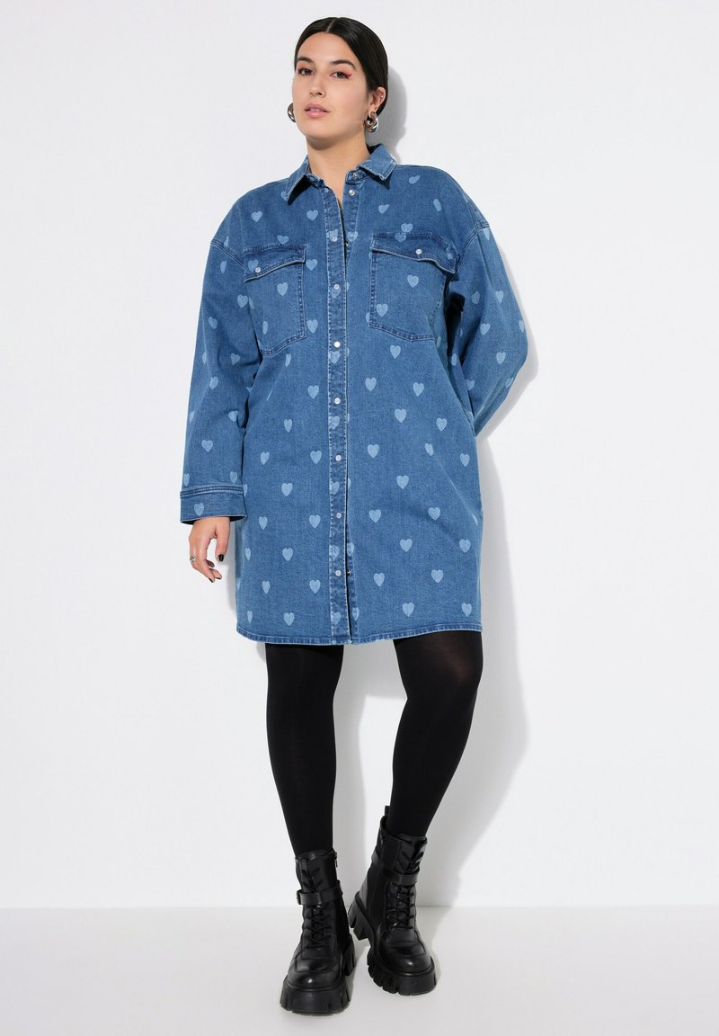 Jeanshemdkleid in Hellblau mit Herzmustern, verfügt über eine Knopfleiste, zwei Brusttaschen und lange Ärmel. Kombiniert mit schwarzen Leggings und klobigen Stiefeln.