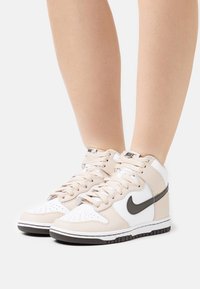 Nike high-top sneakers featuring a white leather base with beige accents, textured details, and a black swoosh. Rubber sole with tread pattern.