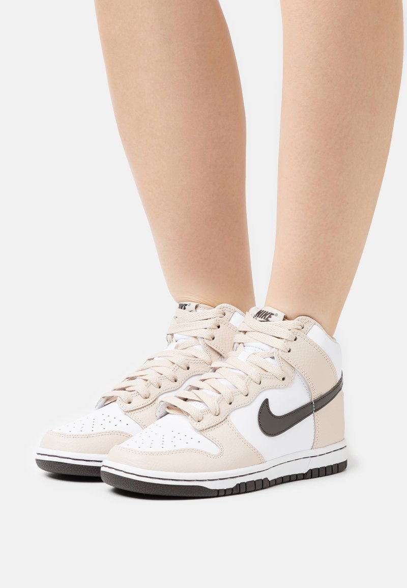 Nike high-top sneakers featuring a white leather base with beige accents, textured details, and a black swoosh. Rubber sole with tread pattern.