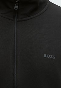 Zwarte jas met rits, hoge kraag en gegraveerd "BOSS"-logo op de linkerborst.