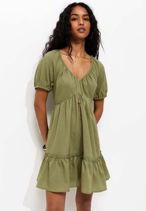 Donna che indossa un vestito verde oliva a maniche corte con orlo arricciato e scollo a V, accessoriato con una lunga collana dorata e orecchini a cerchio.
