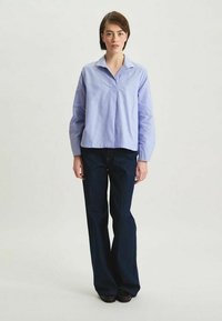 Chemise mauve à manches longues avec col et pli avant, associée à un jean évasé bleu foncé. Texture lisse, coupe décontractée.
