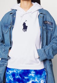 Vit hoodie med en marinblå broderad logotyp av en häst och ryttare, lager på lager under en jeansjacka med en lapp. Tie-dye blå sweatpants.