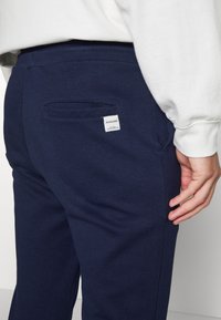 Pantalones de chándal azul marino en una tela suave, con un bolsillo trasero de abertura horizontal y una etiqueta de marca blanca cosida.