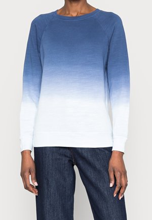 Personne portant un sweat-shirt à manches longues avec un col rond dégradé du bleu au blanc et un jean bleu foncé.