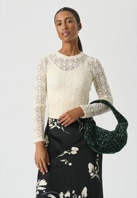 Vrouw in een roomkleurige kanttop en een zwarte bloemenrok, die een donkergroene getextureerde handtas onder haar arm houdt.