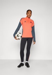 Bright coral and dark gray long-sleeve athletic top with quarter-zip, paired with black leggings featuring red side accents. Holding a soccer ball.