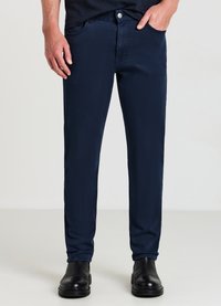 Marineblaue Baumwollmischungshose mit einer figurbetonten Passform, Fünf-Pocket-Design, Knopfverschluss und glatter Textur. Getragen mit schwarzen Lederstiefeln.