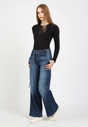 Young woman standing, wearing black long-sleeve top, high-waisted wide-leg blue jeans with drawstring, and white sneakers against a plain background.