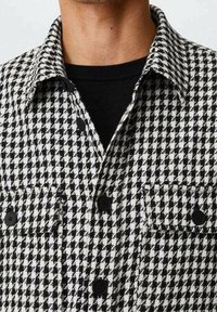 Chemise boutonnée à motif pied-de-poule noir et blanc portée par-dessus un haut noir à col rond, visible du col jusqu'au milieu du torse.