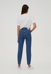 Jean en denim bleu taille haute, coupe slim, ourlets effilochés et deux poches arrière, porté avec un t-shirt blanc uni et des chaussures blanches.