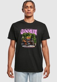 Sort bomulds T-shirt med et grafisk tryk af et hjemme-studie med planter. Lys pink "COOKIN" tekst ovenfor designet.
