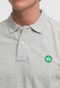 Lys grå polo skjorte laget av teksturert stoff, med en tre-knapps placket og en grønn brodert logo på venstre bryst.