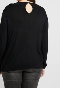 Schwarzes langärmliges Oberteil mit einem Rückenausschnitt. Weicher Stoff, lockere Passform und abgerundeter Saum. Kombiniert mit grauen Jeans für einen lässigen Look.
