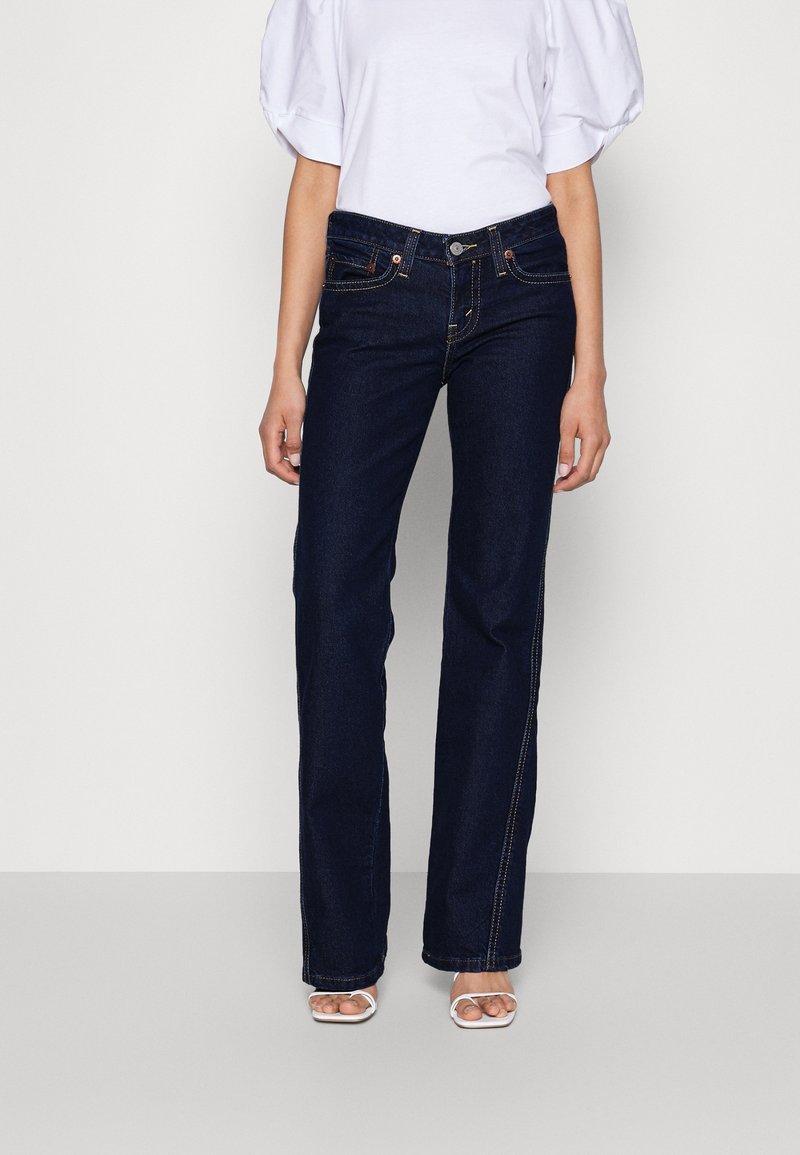 Femme portant un jean bleu foncé ajusté à jambes droites, un chemisier blanc à manches bouffantes et des sandales blanches à talons ouverts, debout devant un fond uni.