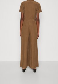 Brun linne jumpsuit med korta ärmar, vida ben, bälte i midjan och slätt, texturerat tyg. Svarta klackskor med blockklack syns.