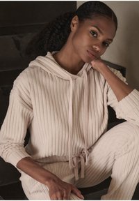 Felpa a coste di un beige chiaro con cappuccio e cordini, abbinata a pantaloni. Texture morbida, vestibilità rilassata, maniche lunghe e design casual e confortevole.
