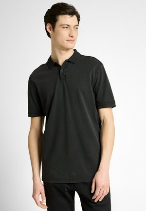 Junger Mann steht, trägt ein schlichtes schwarzes kurzärmeliges Poloshirt und schwarze Hose, schaut leicht nach links vor weißem Hintergrund.