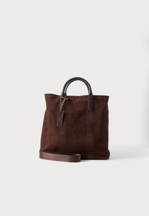 Bolso tote de ante marrón con asas de cuero oscuro, una correa de hombro desmontable y una etiqueta colgante de cuero sobre un fondo blanco.