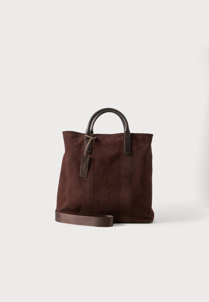 Sac cabas en daim marron avec des poignées en cuir foncé, une bandoulière amovible et une étiquette en cuir suspendue sur fond blanc.