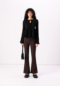 Black textured blouse with a keyhole front, paired with brown flared pants. Model holds a small black handbag and wears chunky black shoes.