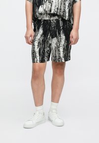 Schwarz-weiß gemusterte Shorts mit elastischem Bund und Seitentaschen, aus leichtem Stoff gefertigt. Kombiniert mit weißen Schuhen und Socken.