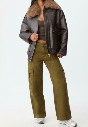 Person trägt eine braune Kunstlederjacke mit Fellkragen, olivgrüne Cargohose, ein kurzes Top und silberne Sneakers.