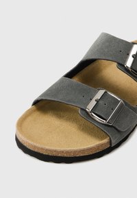 Sandal din suede gri, cu talpă de plută și două catarame argintii. Dispune de un vârf rotund și un design deschis. Solea este din cauciuc texturat.