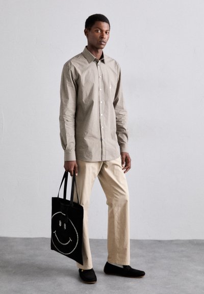 Brun rutig knäppskjorta, beige byxor, svart tote-väska med smileyansikte design, och svarta slip-on skor. Slät tyg och avslappnad passform.