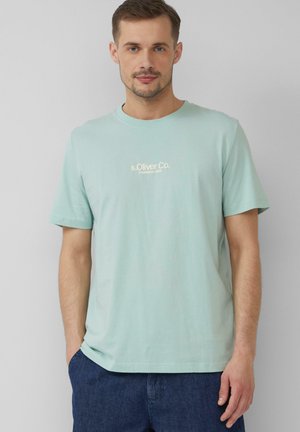 T-shirt en coton teal clair avec un col rond. Présente un petit logo crème sur la poitrine. Manches courtes et coupe décontractée.