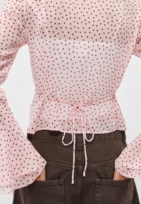 Ανοιχτό ροζ διαφανές μπλουζάκι με σκούρες κόκκινες πουά, μανίκια που φουσκώνουν, σφηκοφωλιά στη μέση με κορδόνι και ποδόγυρο με βολάν.