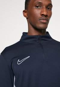 Mørkeblå Nike pullover med lynlås lavet af glat stof, med en stående krave og hvidt logo på venstre bryst samt kontrasterende sideaksenter.
