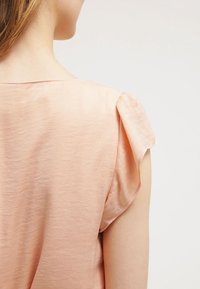 Top de color melocotón con detalles de hombros suavemente plegados y una tela suave y ligera. El diseño presenta un corte relajado y un escote sencillo.
