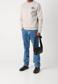 Light gray sweatshirt with ribbed cuffs and embroidered logo, paired with blue denim jeans, black shoulder bag, and black shoes.