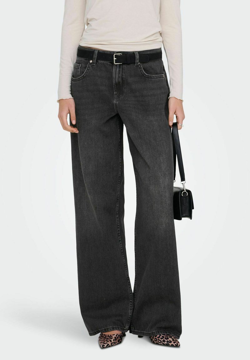 Personne portant un jean noir à jambes larges avec une ceinture noire, tenant un petit sac à main noir et portant des chaussures pointues imprimées léopard.