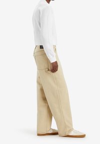 Pantalones de pierna ancha beige con cintura alta, confeccionados en denim. Presentan costuras visibles, bolsillos traseros y una etiqueta negra en la cinturilla.