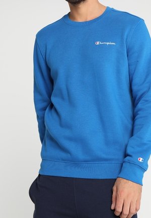 Homme portant un sweat-shirt Champion bleu avec un logo sur la poitrine et la manche, debout devant un fond clair uni.
